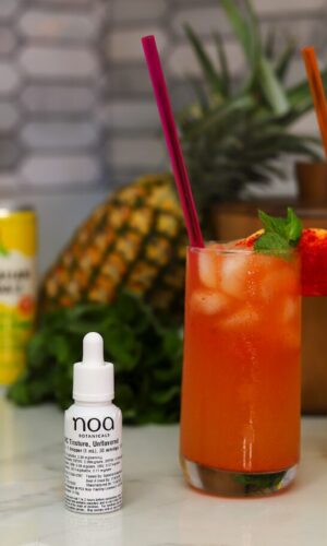 Two colorful cocktails with garnish sit on the counter, alongside a a cannabis THC Nano Tincutre A can of pineapple drink and a fresh pineapple add a tropical touch to the vibrant setting.